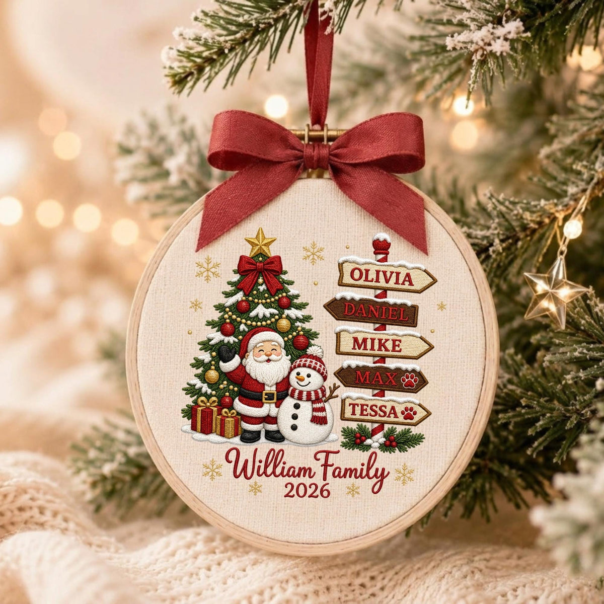 May include: A round wooden embroidery hoop ornament featuring a cream fabric background with a printed Christmas tree, Santa Claus, and a snowman. A red ribbon bow sits at the top. The text reads William Family 2026 and lists Olivia, Daniel, Mike, Max, and Tessa.