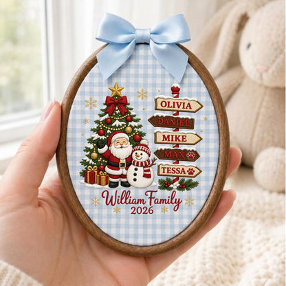 May include: An oval wooden embroidery hoop ornament featuring a light blue gingham fabric background. It displays a Christmas tree, Santa Claus, a snowman, and a signpost with the names Olivia, Daniel, Mike, Max, and Tessa. Text reads William Family 2026, topped with a light blue satin bow.