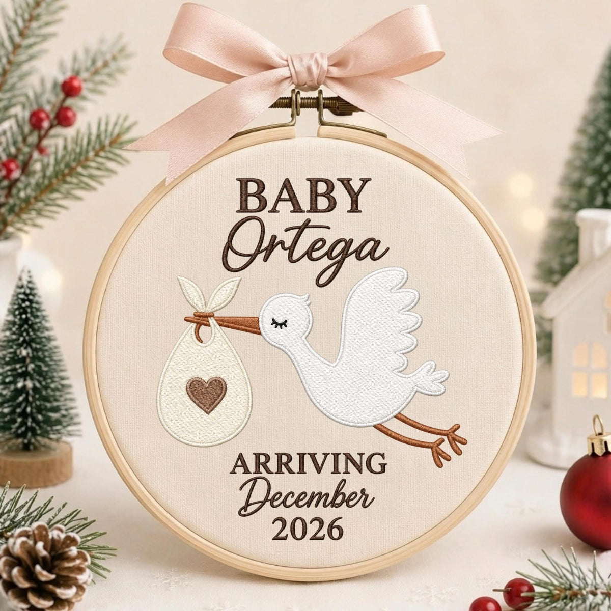 May include: A round wooden embroidery hoop containing cream-colored fabric with embroidered text reading BABY Ortega ARRIVING December 2026. A white stork carries a bundle with a brown heart, topped with a light pink satin ribbon bow.