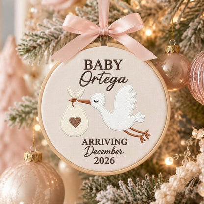 May include: A round wooden embroidery hoop ornament featuring a white fabric center with an embroidered white stork carrying a bundle with a brown heart. The text reads BABY Ortega ARRIVING December 2026. A light pink satin ribbon bow is at the top.