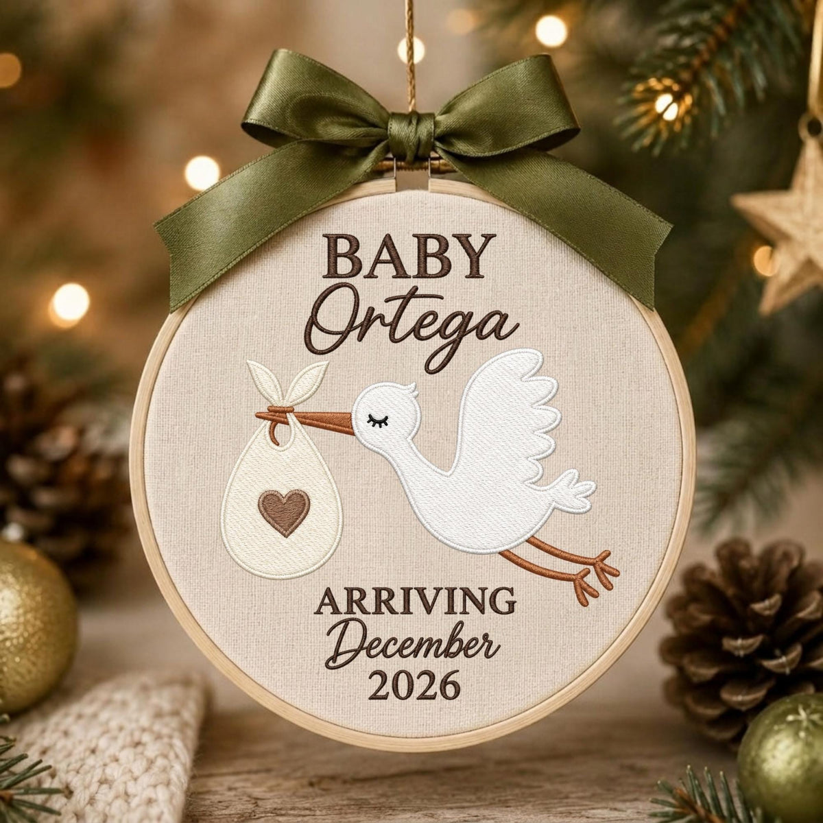 May include: A round embroidery hoop ornament featuring a white stork carrying a bundle with a brown heart. The text reads BABY Ortega ARRIVING December 2026 in brown lettering. An olive green satin ribbon bow is tied at the top of the wooden frame.