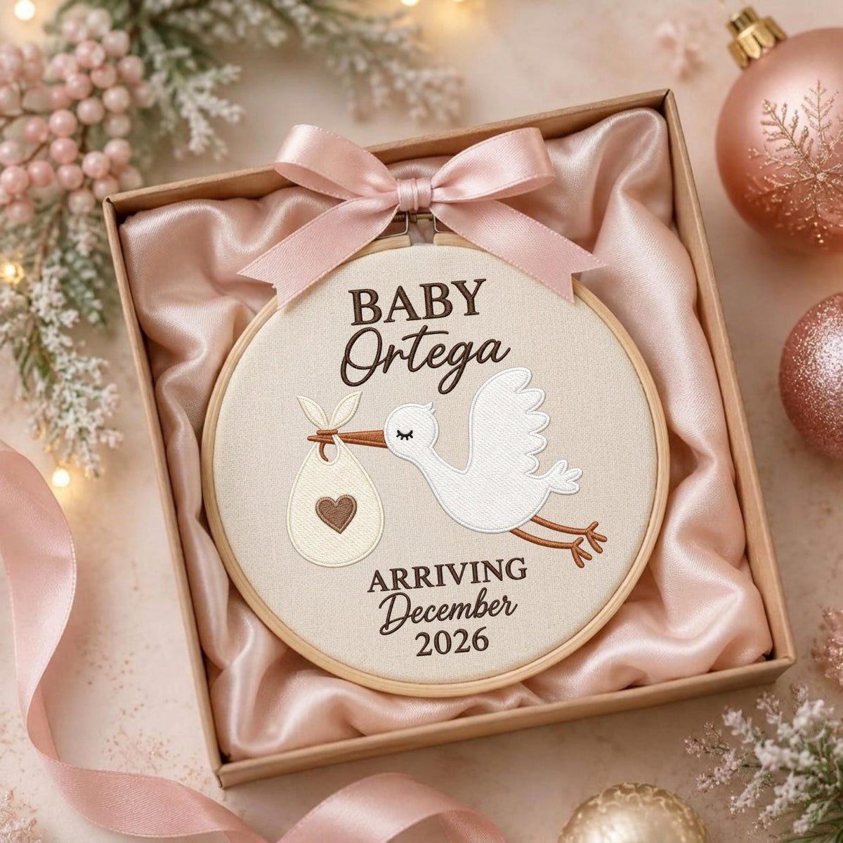 May include: A round wooden embroidery hoop containing beige fabric with an embroidered white stork carrying a bundle with a brown heart. The text reads BABY Ortega ARRIVING December 2026. A light pink satin ribbon bow is attached to the top.