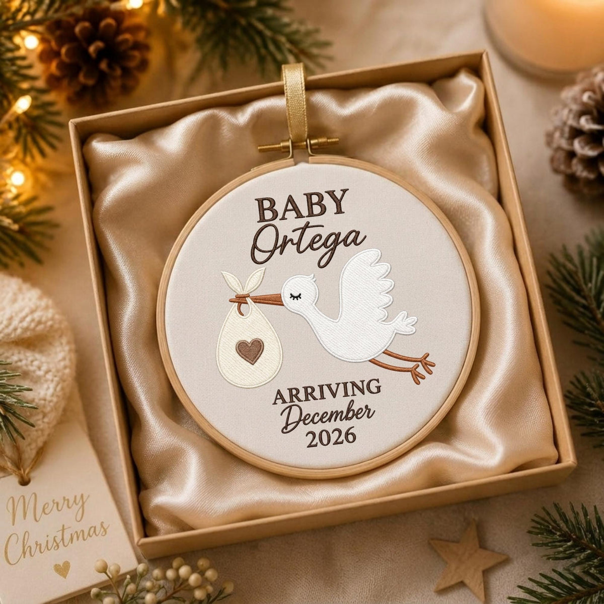 May include: A round embroidery hoop ornament featuring a white stork carrying a bundle with a brown heart. The text reads BABY Ortega ARRIVING December 2026. The ornament is presented in a square box lined with beige satin fabric and a gold ribbon.