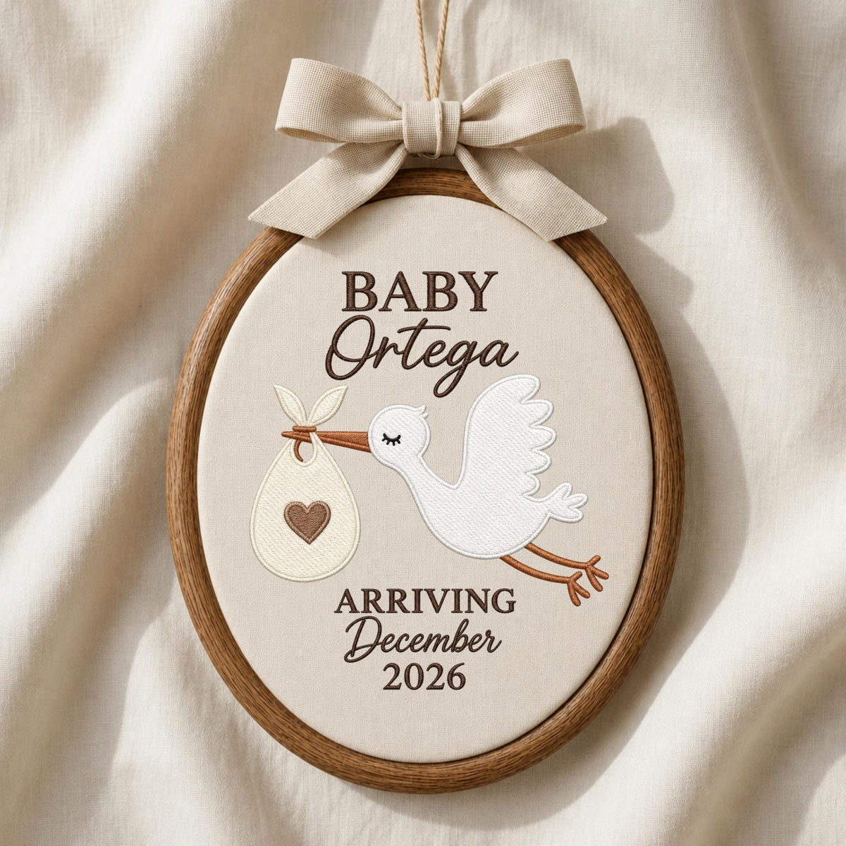 May include: An oval embroidery hoop featuring a cream-colored fabric with a brown embroidered stork carrying a bundle with a heart. The text reads BABY Ortega ARRIVING December 2026. A beige fabric bow is attached to the top of the wooden frame.