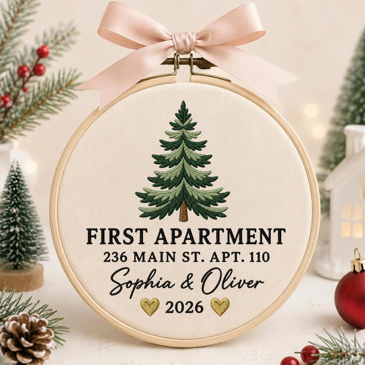May include: A round wooden embroidery hoop containing cream-colored fabric with an embroidered green pine tree. The text reads FIRST APARTMENT, 236 MAIN ST. APT. 110, Sophia &amp; Oliver, and 2026, with two small gold hearts. A light pink ribbon bow is attached.