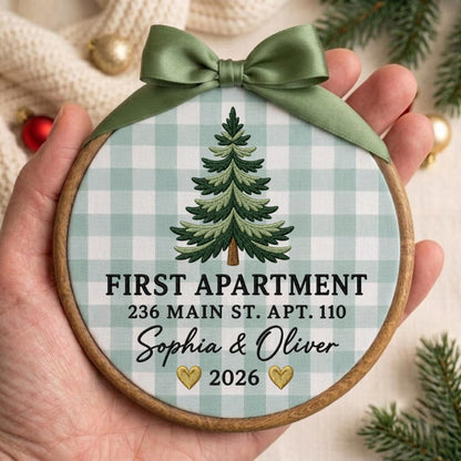 May include: A round wooden embroidery hoop ornament featuring a light green and white gingham fabric background. A green embroidered Christmas tree sits above the text FIRST APARTMENT 236 MAIN ST. APT. 110, Sophia &amp; Oliver, and 2026 with two gold hearts.