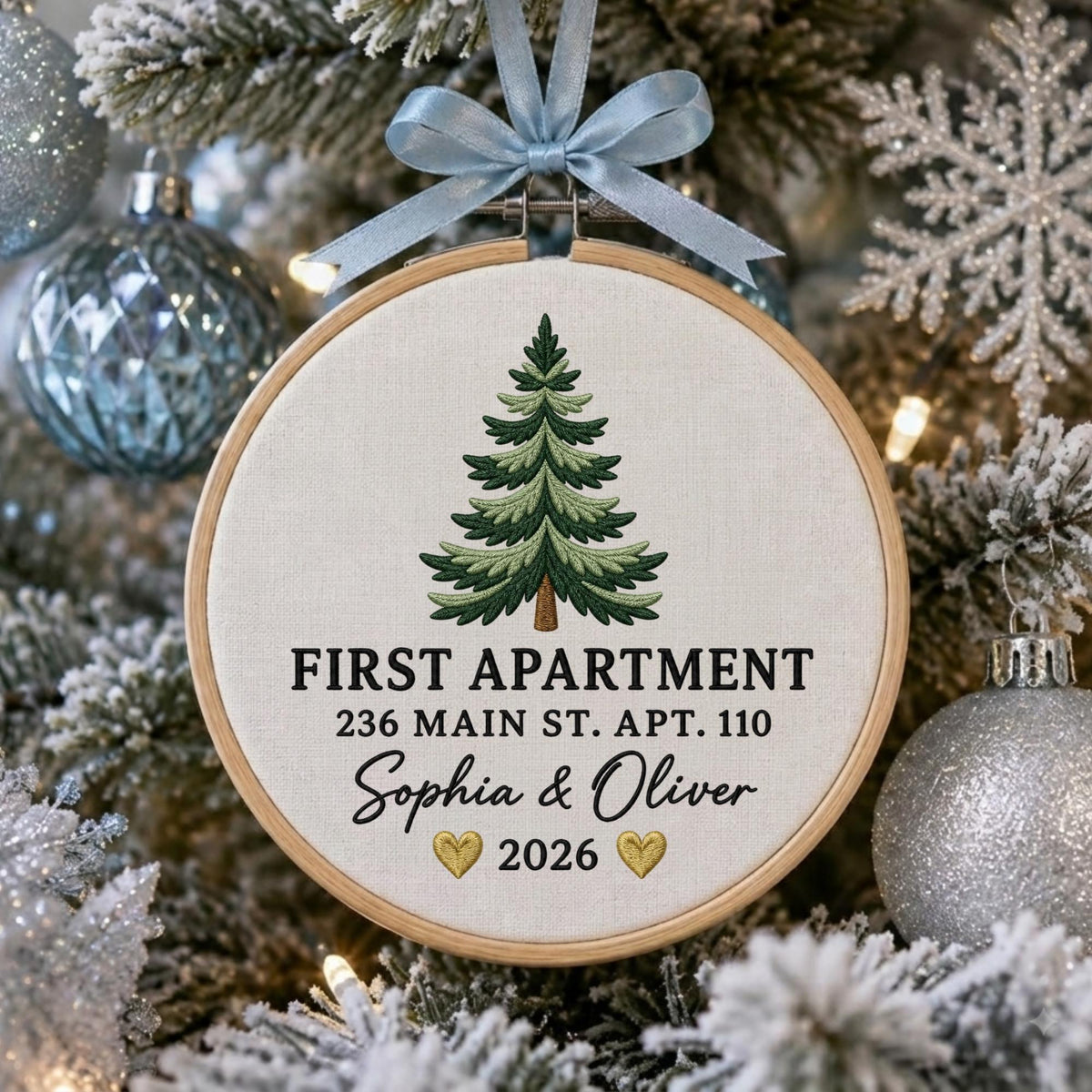 May include: A round wooden embroidery hoop ornament featuring a light-colored fabric background with an embroidered green pine tree. The text reads FIRST APARTMENT, 236 MAIN ST. APT. 110, Sophia &amp; Oliver, and 2026 with two small gold hearts.