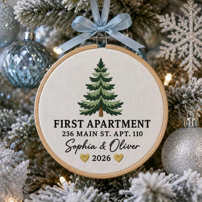 May include: A round wooden embroidery hoop ornament featuring a light-colored fabric background with an embroidered green pine tree. The text reads FIRST APARTMENT, 236 MAIN ST. APT. 110, Sophia &amp; Oliver, and 2026 with two small gold hearts.