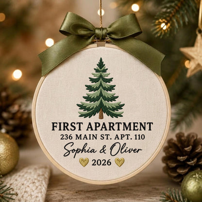 May include: A round wooden embroidery hoop ornament featuring a cream-colored fabric center with an embroidered green pine tree. Text reads FIRST APARTMENT 236 MAIN ST. APT. 110, Sophia &amp; Oliver, and 2026 with two gold hearts. An olive green satin ribbon bow is tied at the top.