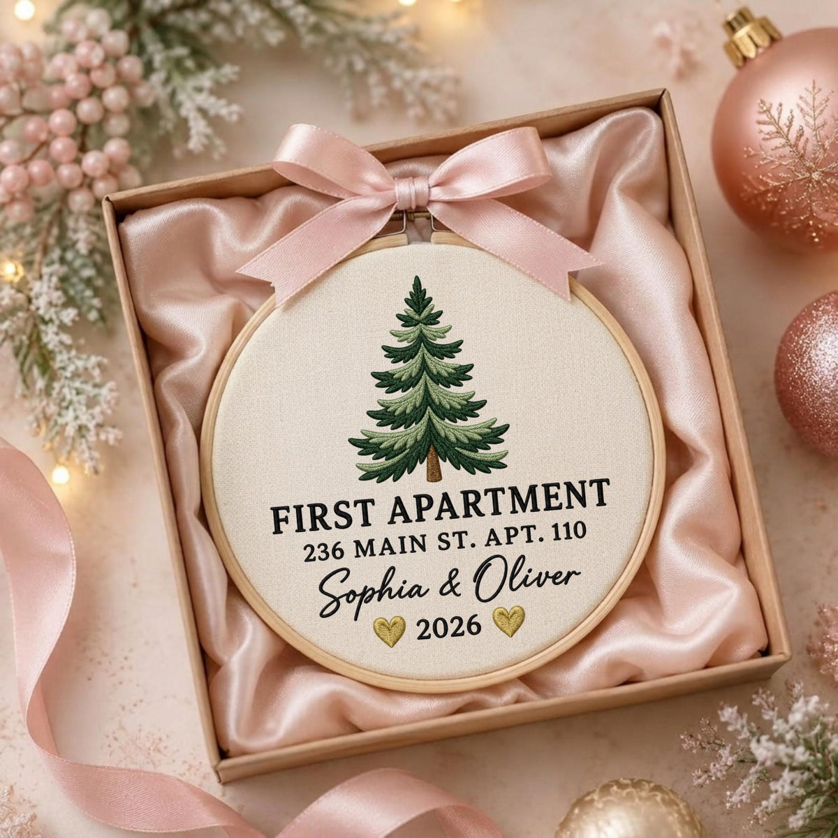 May include: A round wooden embroidery hoop containing cream fabric with a green embroidered pine tree. The text reads FIRST APARTMENT 236 MAIN ST. APT. 110 Sophia &amp; Oliver 2026 with two small gold hearts. A light pink satin ribbon bow is attached.