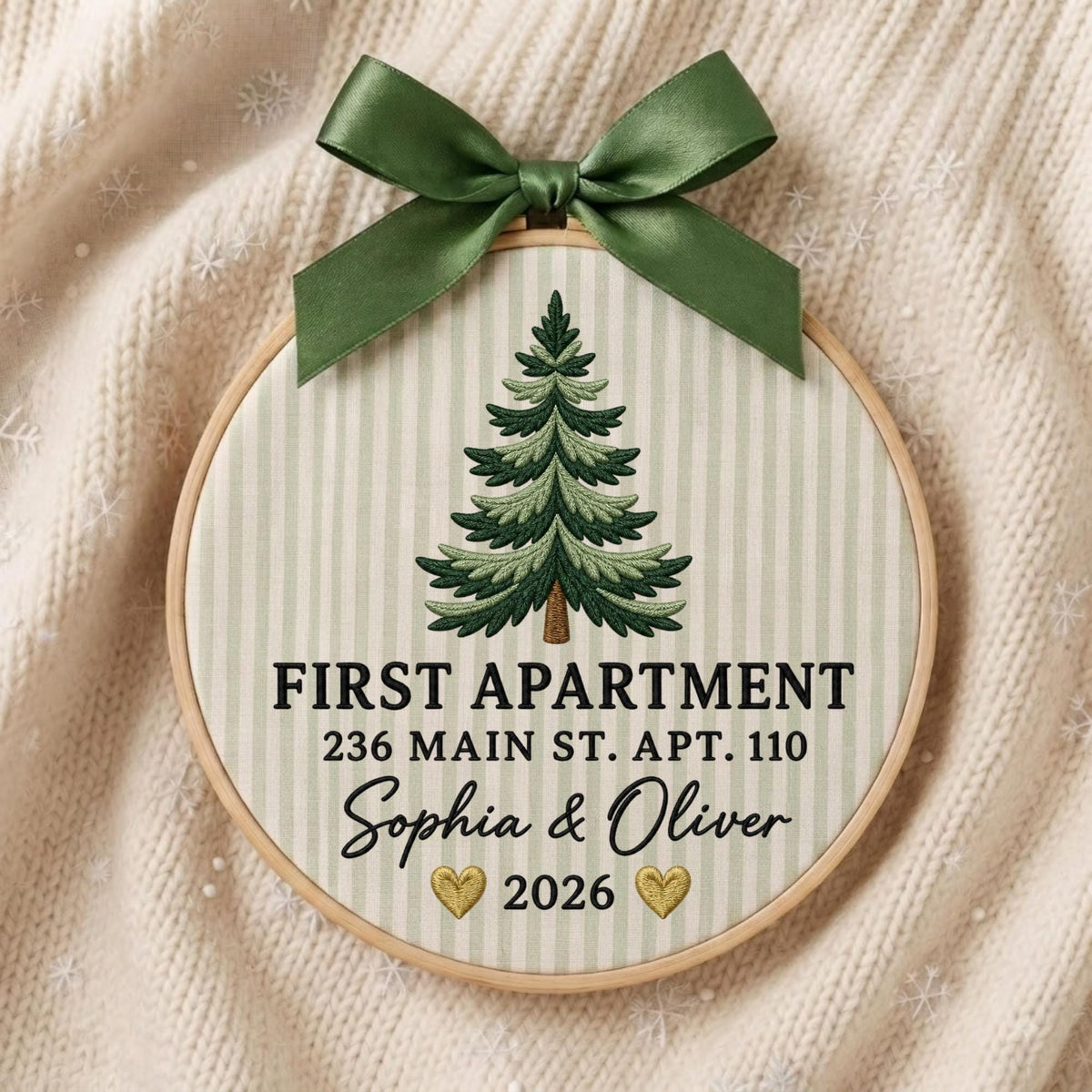 May include: A circular embroidery hoop featuring a green pine tree design on a light green striped fabric. The text reads FIRST APARTMENT, 236 MAIN ST. APT. 110, Sophia &amp; Oliver, and 2026 with two gold hearts. A dark green satin ribbon bow is attached.