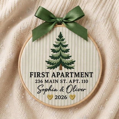 May include: A circular embroidery hoop featuring a green pine tree design on a light green striped fabric. The text reads FIRST APARTMENT, 236 MAIN ST. APT. 110, Sophia &amp; Oliver, and 2026 with two gold hearts. A dark green satin ribbon bow is attached.