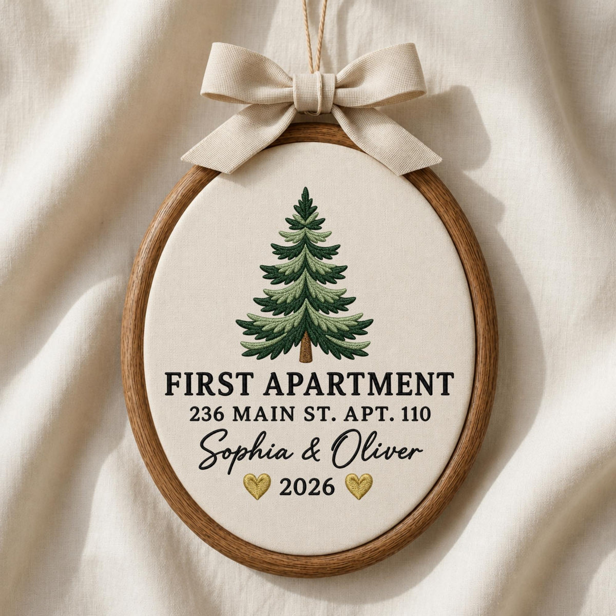 May include: An oval wooden embroidery hoop ornament featuring a cream-colored fabric background with an embroidered green pine tree. The text reads FIRST APARTMENT 236 MAIN ST. APT. 110 Sophia &amp; Oliver 2026 with two small gold hearts and a beige ribbon bow.