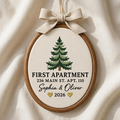 May include: An oval wooden embroidery hoop ornament featuring a cream-colored fabric background with an embroidered green pine tree. The text reads FIRST APARTMENT 236 MAIN ST. APT. 110 Sophia &amp; Oliver 2026 with two small gold hearts and a beige ribbon bow.