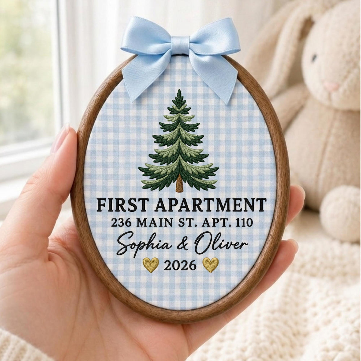 May include: An oval wooden embroidery hoop frame containing fabric with a light blue gingham pattern. It features an embroidered green pine tree, the text FIRST APARTMENT 236 MAIN ST. APT. 110, the names Sophia &amp; Oliver, and the year 2026 with two gold hearts, topped with a light blue satin ribbon bow.