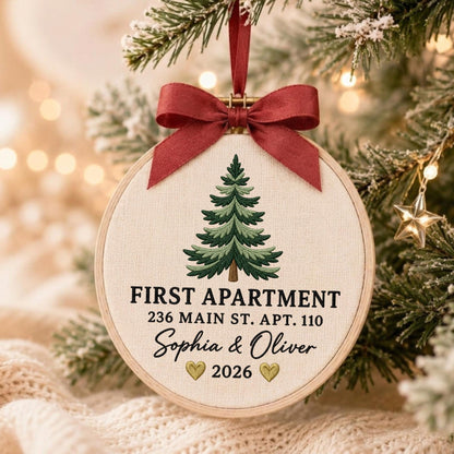 May include: A round wooden embroidery hoop ornament featuring a cream-colored fabric center with an embroidered green pine tree. The text reads FIRST APARTMENT, 236 MAIN ST. APT. 110, Sophia &amp; Oliver, and 2026 with two small gold hearts. A dark red ribbon bow is attached at the top.