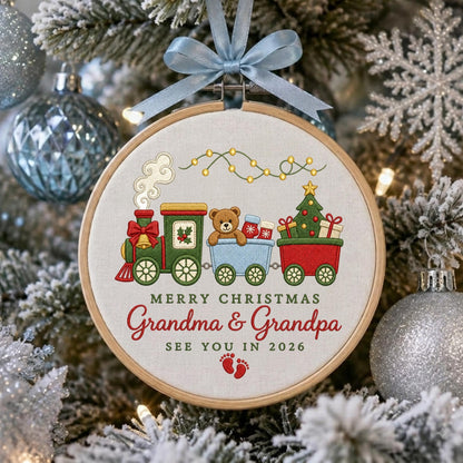 May include: A round wooden embroidery hoop ornament featuring a stitched Christmas train with a teddy bear, a decorated tree, and gifts. The text reads MERRY CHRISTMAS GRANDMA &amp; GRANDPA SEE YOU IN 2026 with red baby footprints. A light blue ribbon bow is attached.