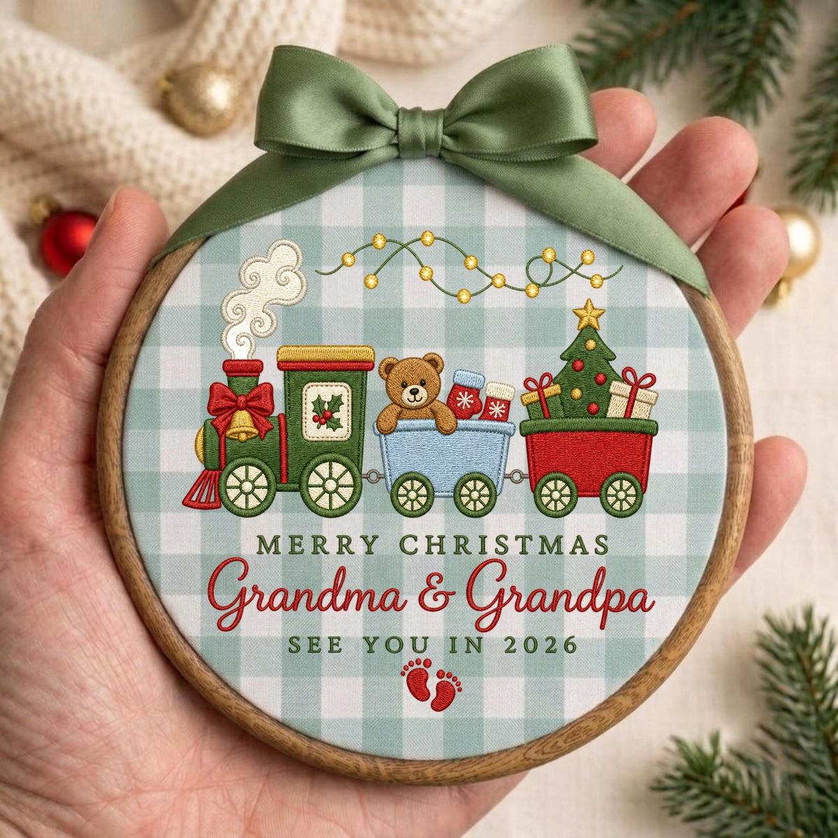 May include: A round wooden embroidery hoop ornament featuring a light blue and white buffalo check fabric. The design shows a green and red train carrying a teddy bear and a Christmas tree, with the text Merry Christmas Grandma &amp; Grandpa See You In 2026 and red baby footprints, topped with a sage green satin ribbon bow.