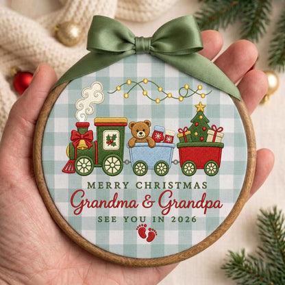 May include: A round wooden embroidery hoop ornament featuring a light blue and white buffalo check fabric. The design shows a green and red train carrying a teddy bear and a Christmas tree, with the text Merry Christmas Grandma &amp; Grandpa See You In 2026 and red baby footprints, topped with a sage green satin ribbon bow.