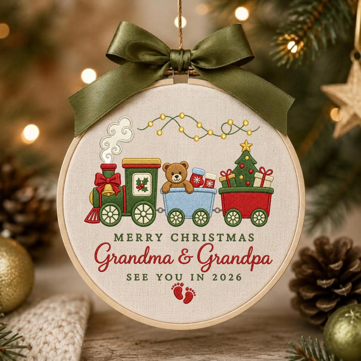May include: A round embroidery hoop ornament featuring a green train with a teddy bear, a Christmas tree, and gifts. The text reads MERRY CHRISTMAS Grandma &amp; Grandpa SEE YOU IN 2026 with red baby footprints. An olive green satin bow tops the hoop.