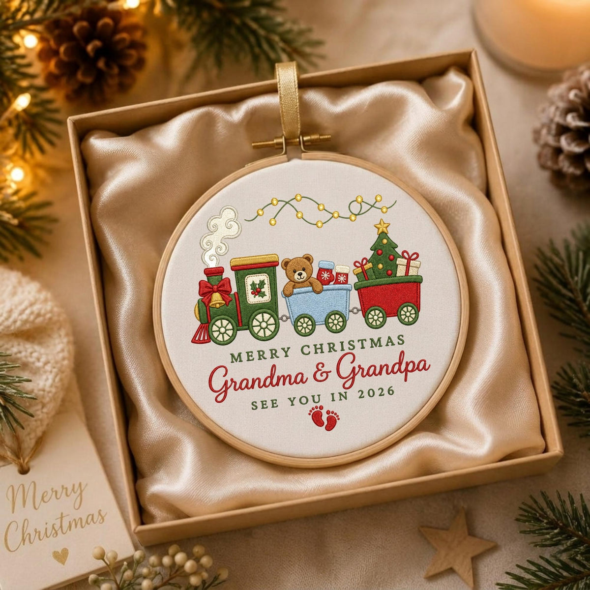 May include: A circular embroidery hoop ornament featuring a stitched Christmas train with a teddy bear, a decorated tree, and gifts. The text reads Merry Christmas Grandma &amp; Grandpa See You In 2026 with two red baby footprints below the lettering.