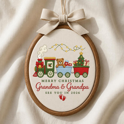 May include: An oval embroidery hoop ornament featuring a cream fabric background with a festive train design. The train carries a teddy bear and a Christmas tree. Text reads: MERRY CHRISTMAS Grandma &amp; Grandpa SEE YOU IN 2026, with red baby footprints.