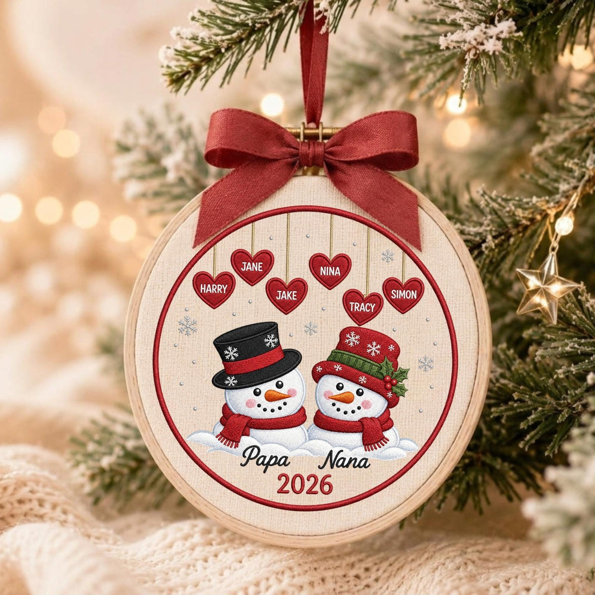 May include: A round embroidery hoop ornament featuring two snowmen wearing hats and scarves, with six hanging red hearts labeled Harry, Jane, Jake, Nina, Tracy, and Simon. The text Papa Nana 2026 is printed below, topped with a dark red ribbon bow.