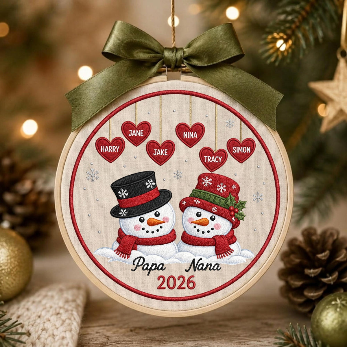 May include: A round wooden embroidery hoop ornament featuring two embroidered snowmen with red scarves and hats. Above them, six red hearts hang with the names Harry, Jane, Jake, Nina, Tracy, and Simon. The text Papa Nana 2026 is below, topped with an olive green ribbon.