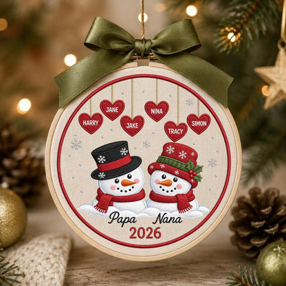 May include: A round wooden embroidery hoop ornament featuring two embroidered snowmen with red scarves and hats. Above them, six red hearts hang with the names Harry, Jane, Jake, Nina, Tracy, and Simon. The text Papa Nana 2026 is below, topped with an olive green ribbon.
