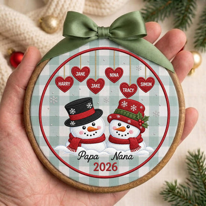 May include: A round wooden embroidery hoop ornament featuring a light green and white checkered fabric background. The design shows two snowmen, one in a black top hat and one in a red hat, with hanging red hearts labeled Harry, Jane, Jake, Nina, Tracy, Simon, and the text Papa Nana 2026. An olive green satin ribbon bow is attached to the top.