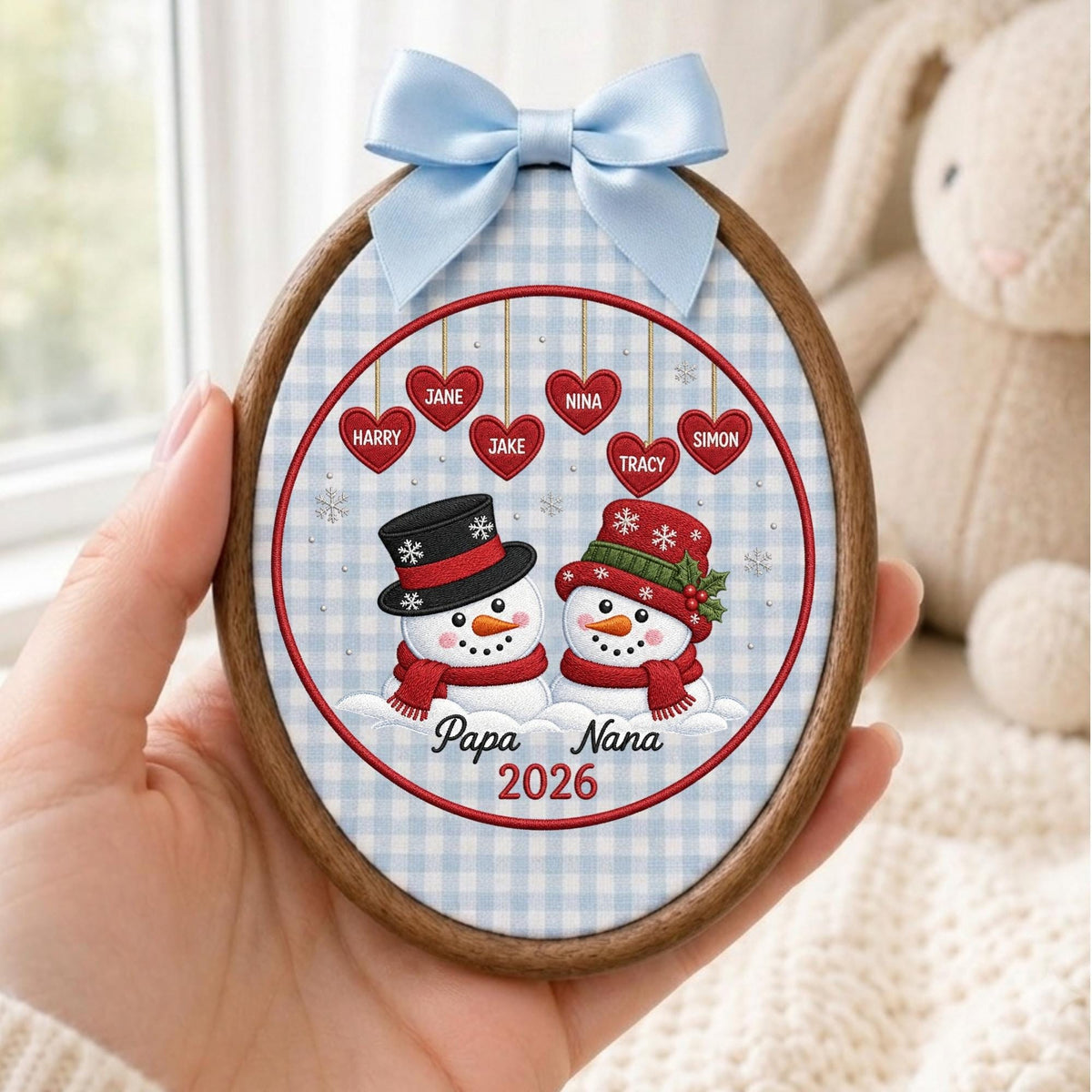 May include: An oval wooden embroidery hoop frame containing fabric with a light blue gingham pattern. It features two snowmen, one with a black top hat and one with a red hat, below six red hearts with names and the text Papa Nana 2026.
