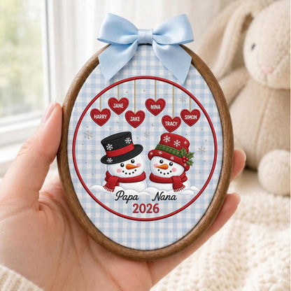May include: An oval wooden embroidery hoop frame containing fabric with a light blue gingham pattern. It features two snowmen, one with a black top hat and one with a red hat, below six red hearts with names and the text Papa Nana 2026.