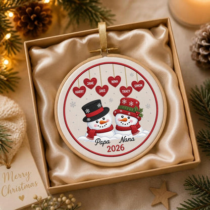 May include: A round embroidery hoop ornament featuring two snowmen wearing hats and scarves. Above them, six red hearts hang by strings, each inscribed with a name. The text Papa Nana 2026 is printed below the snowmen. The ornament is in a gift box.