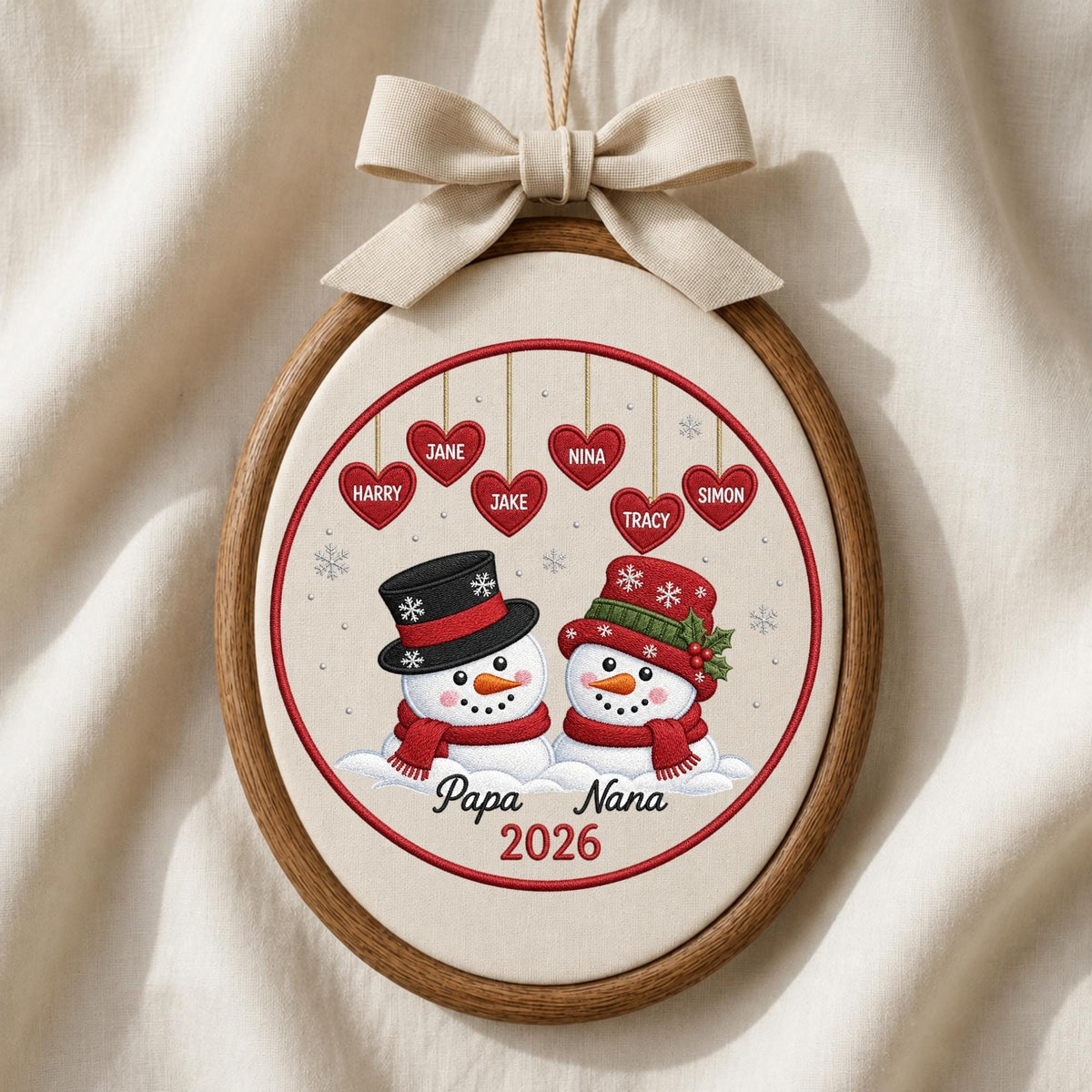 May include: An oval wooden embroidery hoop ornament featuring two snowmen with red scarves and hats. Above them, six red hearts hang with the names Harry, Jane, Jake, Nina, Tracy, and Simon. The text Papa Nana 2026 is printed below, topped with a beige bow.