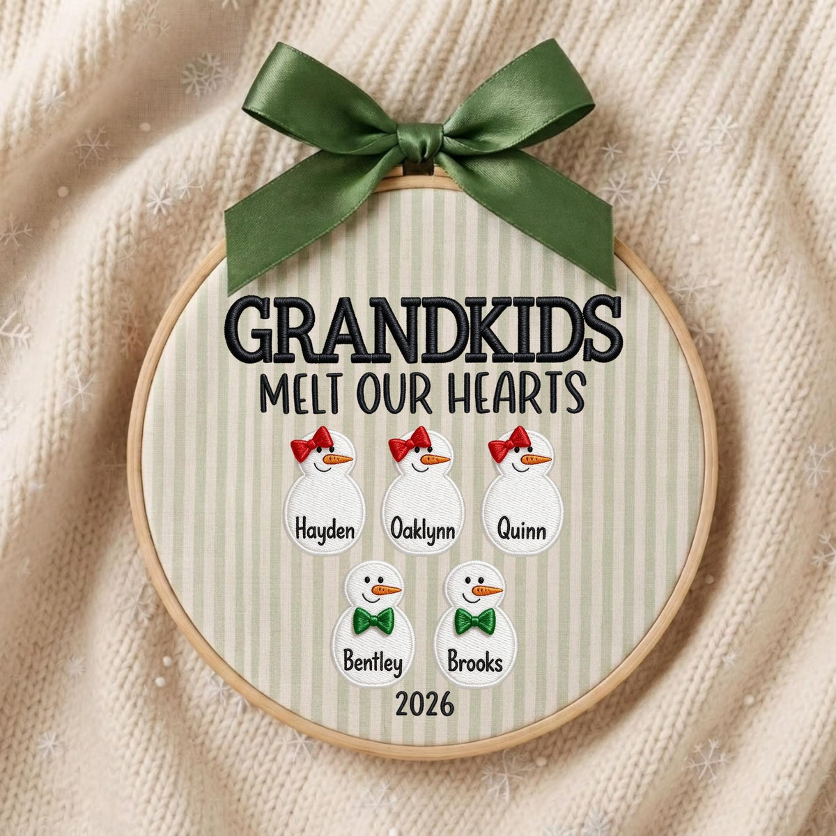 May include: A circular wooden embroidery hoop frame containing fabric with vertical light green stripes. The text "GRANDKIDS MELT OUR HEARTS" is embroidered in black above five snowman figures with names "Hayden", "Oaklynn", "Quinn", "Bentley", "Brooks" and "2026". A dark green ribbon bow is tied at the top.
