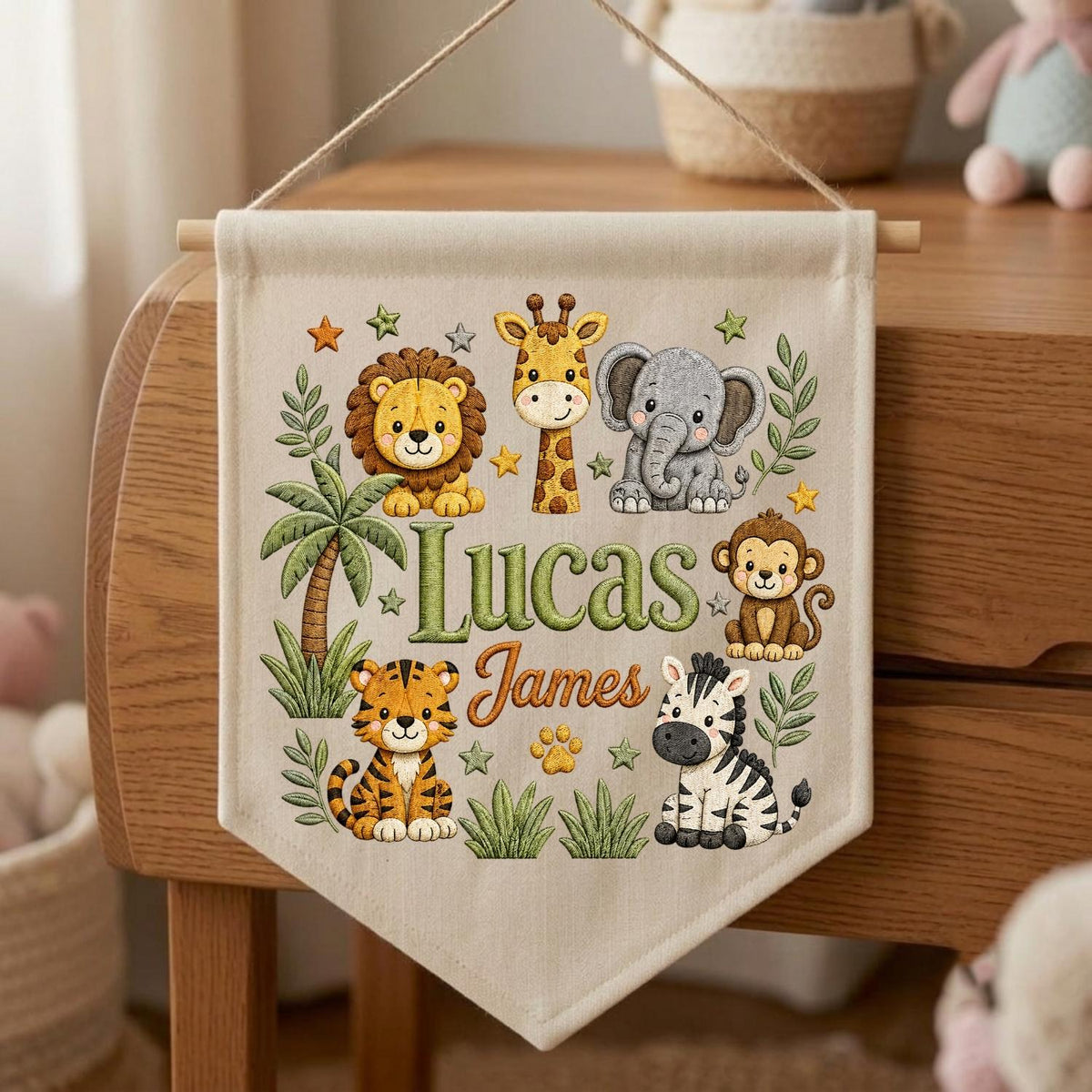 May include: A cream-colored fabric wall banner featuring a hanging string and wooden dowel. The banner displays illustrations of a lion, giraffe, elephant, monkey, tiger, and zebra, along with green palm trees and stars. The text reads Lucas James.