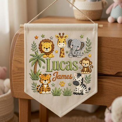 May include: A cream-colored fabric wall banner featuring a hanging string and wooden dowel. The banner displays illustrations of a lion, giraffe, elephant, monkey, tiger, and zebra, along with green palm trees and stars. The text reads Lucas James.