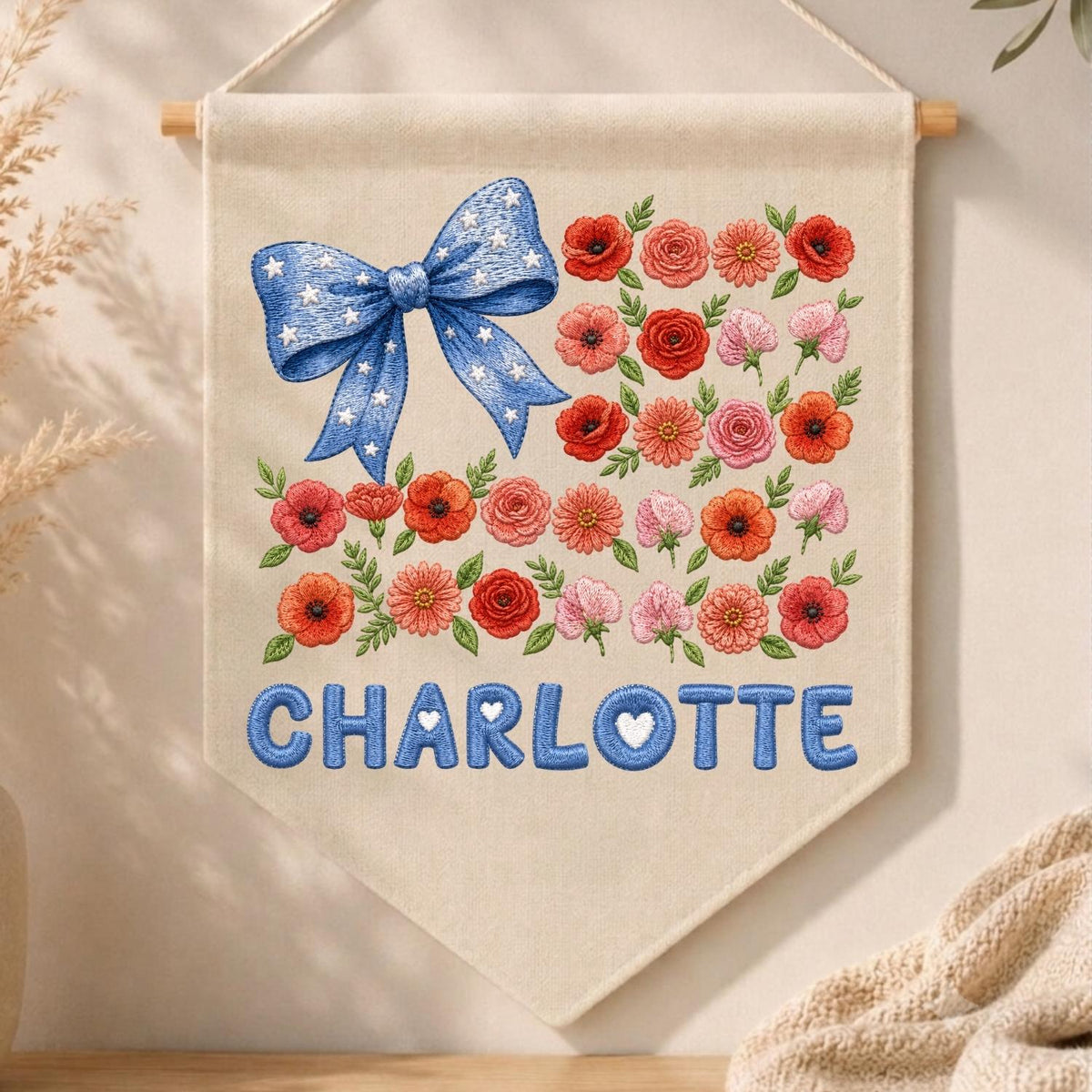 May include: A cream-colored fabric wall banner featuring an embroidered blue bow with white stars in the upper left corner. The design includes various red, orange, and pink flowers arranged in rows. The name CHARLOTTE is embroidered in blue.