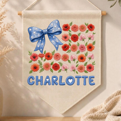 May include: A cream-colored fabric wall banner featuring an embroidered blue bow with white stars in the upper left corner. The design includes various red, orange, and pink flowers arranged in rows. The name CHARLOTTE is embroidered in blue.