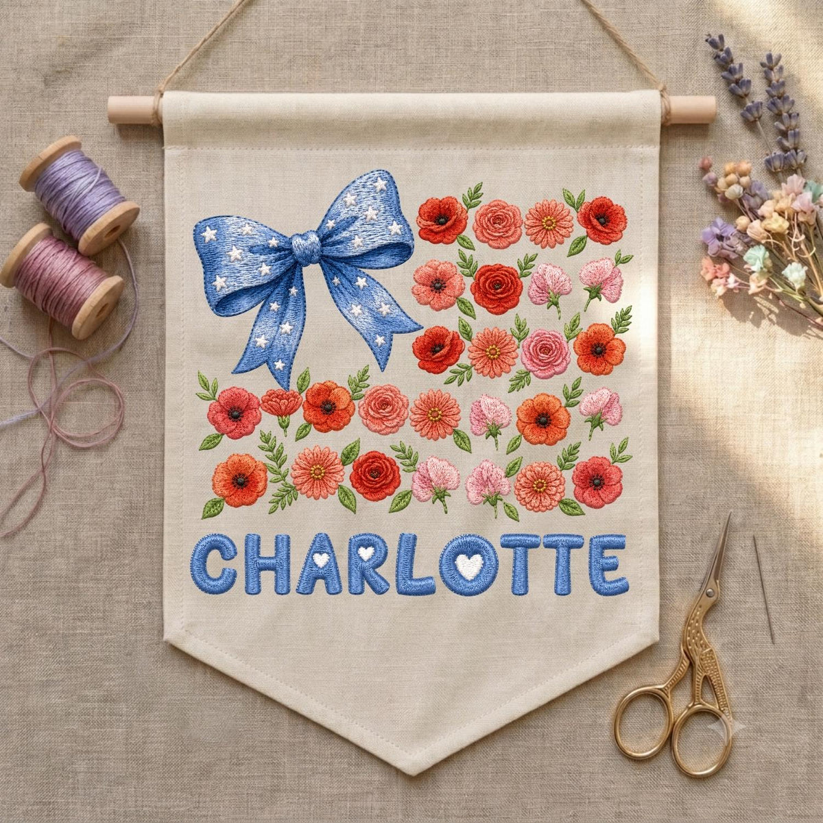 May include: A beige fabric wall banner featuring an embroidered blue bow with white stars, a collection of red, orange, and pink flowers with green leaves, and the name CHARLOTTE in blue letters with small white hearts inside the letters.