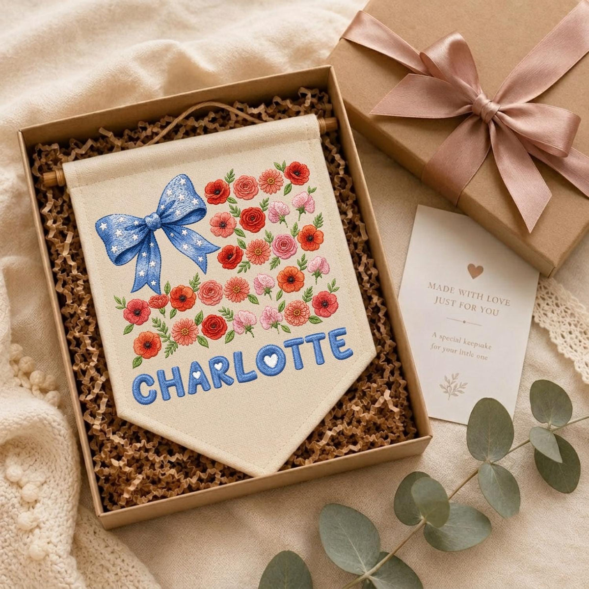 May include: A cream-colored fabric wall banner featuring an embroidered blue bow with white stars, a cluster of red and pink flowers, and the name CHARLOTTE in blue letters, presented in a gift box with a card reading MADE WITH LOVE.