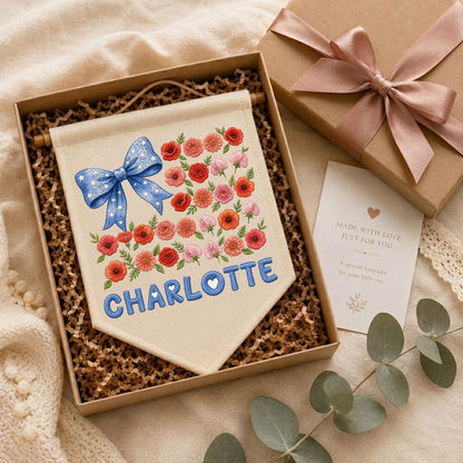 May include: A cream-colored fabric wall banner featuring an embroidered blue bow with white stars, a cluster of red and pink flowers, and the name CHARLOTTE in blue letters, presented in a gift box with a card reading MADE WITH LOVE.