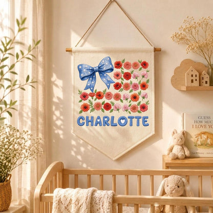 May include: A cream-colored fabric wall hanging banner featuring a blue bow with white stars on the left and a cluster of red, orange, and pink flowers on the right. The name CHARLOTTE is printed in large blue letters at the bottom of the banner.