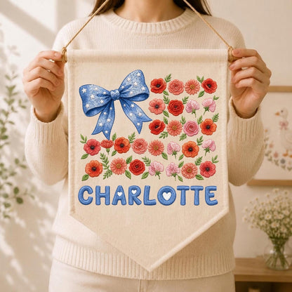 May include: A cream-colored fabric wall banner featuring a large blue bow with white stars, a collection of red and pink floral illustrations, and the name CHARLOTTE printed in blue block letters with a small white heart inside the letter O.