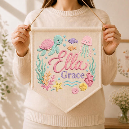 May include: A cream-colored fabric wall banner featuring a colorful underwater scene with a sea turtle, jellyfish, starfish, coral, and shells. The name Ella Grace is printed in the center in a pink script font with purple block lettering.