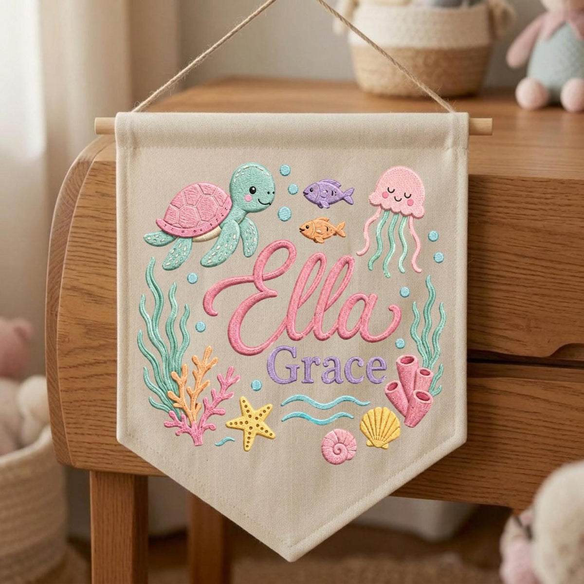 May include: A beige fabric wall banner featuring embroidered sea life including a green turtle, pink jellyfish, orange fish, yellow starfish, and coral. The name Ella Grace is embroidered in the center in pink and purple cursive script lettering.