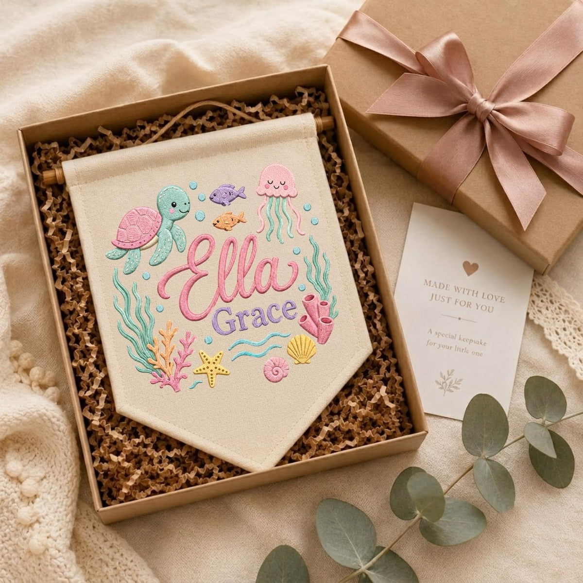 May include: A cream-colored fabric wall banner featuring embroidered sea life including a turtle, jellyfish, starfish, and shells, with the name Ella Grace in pink and purple script. The banner is presented in a gift box with a card.
