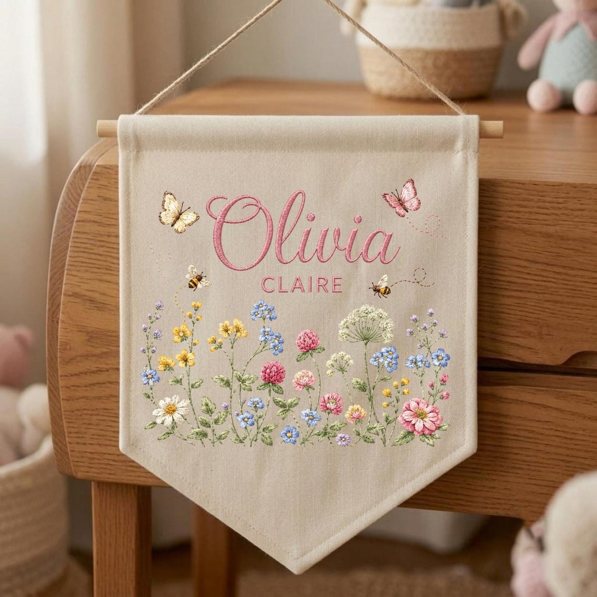 May include: A beige fabric wall banner featuring the embroidered name Olivia Claire in pink script. The design includes colorful wildflowers, two small bees, and two butterflies in shades of yellow, blue, pink, and white, suspended by a wooden rod and string.
