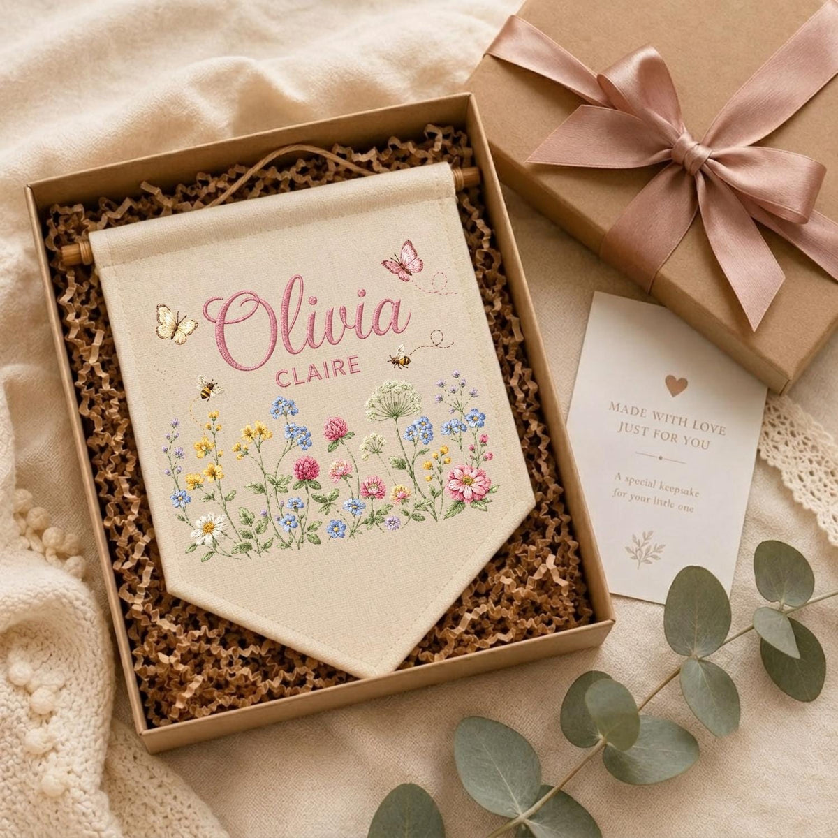 May include: A cream-colored fabric wall banner featuring the name Olivia Claire in pink script and block letters, surrounded by colorful wildflower illustrations, bees, and butterflies, presented in a gift box with a ribbon and a note.