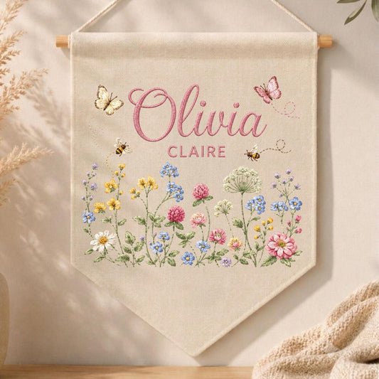 May include: A beige fabric wall banner featuring the embroidered names Olivia and Claire in pink script and block lettering. The design includes colorful wildflowers, green stems, two small bees, and two butterflies in shades of pink and yellow.